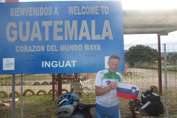 Moški v kolesarski opremi s slovensko zastavo, slikan pred tablo Guatemala.