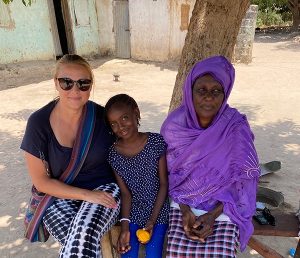 Tri osebe sedijo v senci drevesa: ženska s sončnimi očali, temnopolta deklica in temnopolta starejša ženska v vijoličnem oblačilu