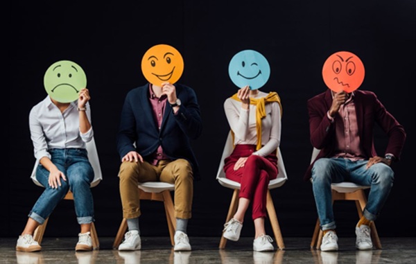 Quattro persone siedono in fila e tengono dei cerchi davanti al viso con diverse espressioni di emozioni: tristezza, gioia, pace e rabbia.