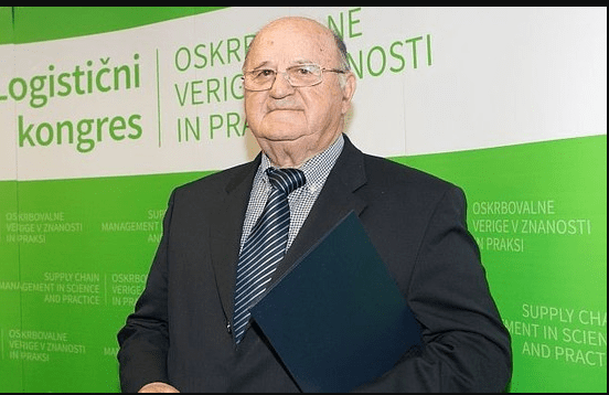 Oseba v temni poslovni obleki in črtasti kravati stoji pred zeleno-belim promocijskim ozadjem logističnega kongresa. V rokah drži temno mapo. Na ozadju so vidni napisi v zelenih odtenkih o logistiki.